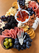 Load image into Gallery viewer, A Halloween-themed charcuterie board arranged on a wooden table, featuring grapes, blackberries, sliced salami, cheese cubes, crackers, almonds, olives, a small skull-shaped cheese, a syringe-shaped dip server, plastic spiders, a skeleton hand, a ghost, and skull-shaped chocolates.