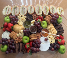 Load image into Gallery viewer, A large charcuterie board arranged on a wooden table, featuring sliced bread, cheeses, crackers, grapes, strawberries, green apples, nuts, and a honey dipper. The board is covered with clear plastic wrap.