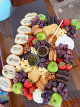 Load image into Gallery viewer, A large charcuterie board arranged on a wooden table, featuring sliced bread, crackers, cheese wedges, grapes, strawberries, green apples, nuts, and two small dipping bowls. A blue textured cup is partially visible at the top left.
