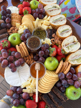 Load image into Gallery viewer, A large charcuterie or cheese board arranged on a tray, featuring sliced bread with spreads, grapes, strawberries, green apples, crackers, cheese wedges, nuts, a small jar of green spread, a jar of dark spread, a honey dipper with honey, and a round cheese wheel.