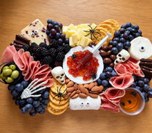 Load image into Gallery viewer, A Halloween-themed charcuterie board arranged on a wooden surface, featuring cheeses, crackers, grapes, salami, almonds, olives, chocolate, and small spooky props like plastic spiders, skull decorations, a skeleton hand, and a syringe garnish.