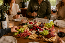 Load image into Gallery viewer, A large charcuterie board filled with cheeses, cured meats, crackers, grapes, nuts, and fruit sits on a wooden dining table, surrounded by people holding wine glasses during a social gathering.